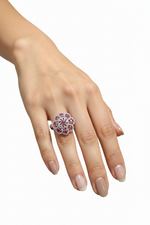 A close-up of a hand wearing a floral cluster ring with pink gemstones and a silver-toned setting, photographed against a plain light gray background.