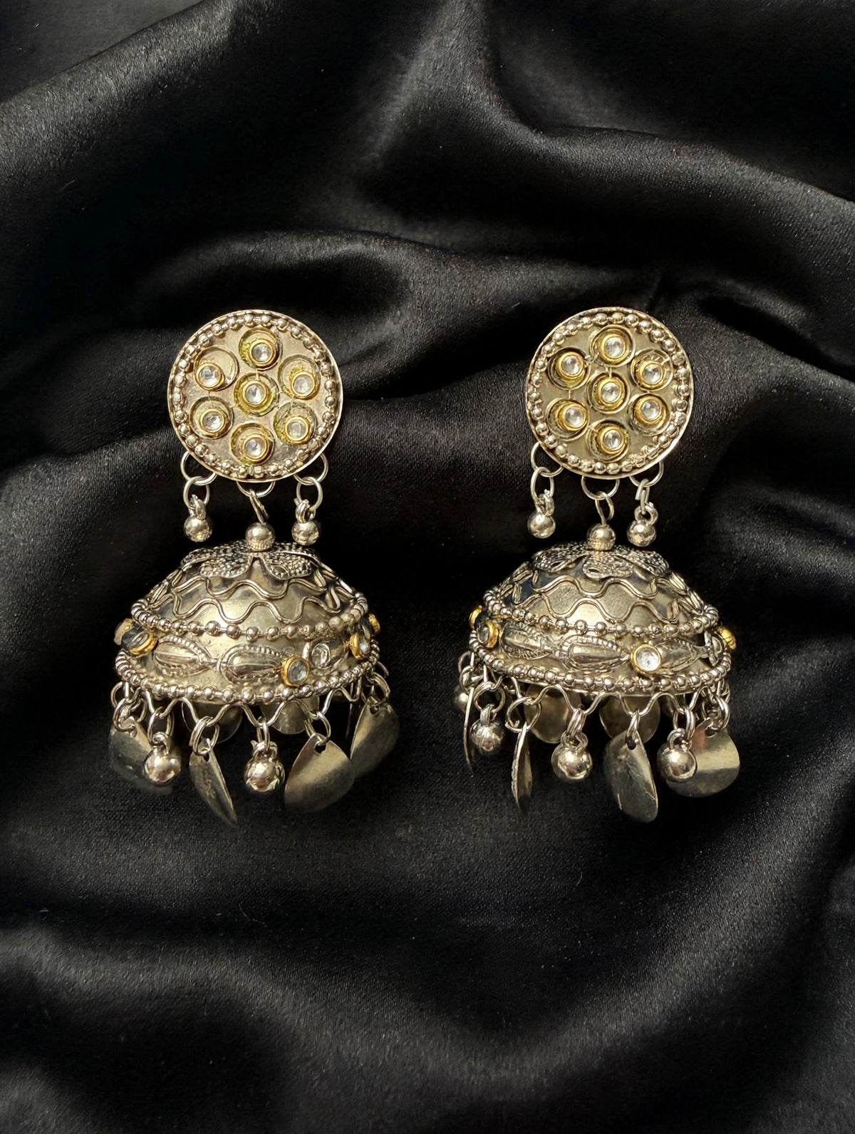 Pair of dual-tone kundan jhumka earrings on black fabric, showing round stone-set tops, floral dome detailing and dangling charms