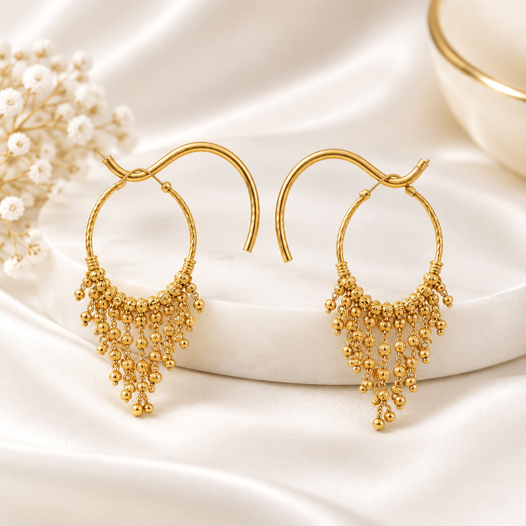 Pair of 22K gold-plated beaded tassel hoop earrings displayed on elegant wooden and gold stand, dark wood interior background — luxury Pakistani jewellry.