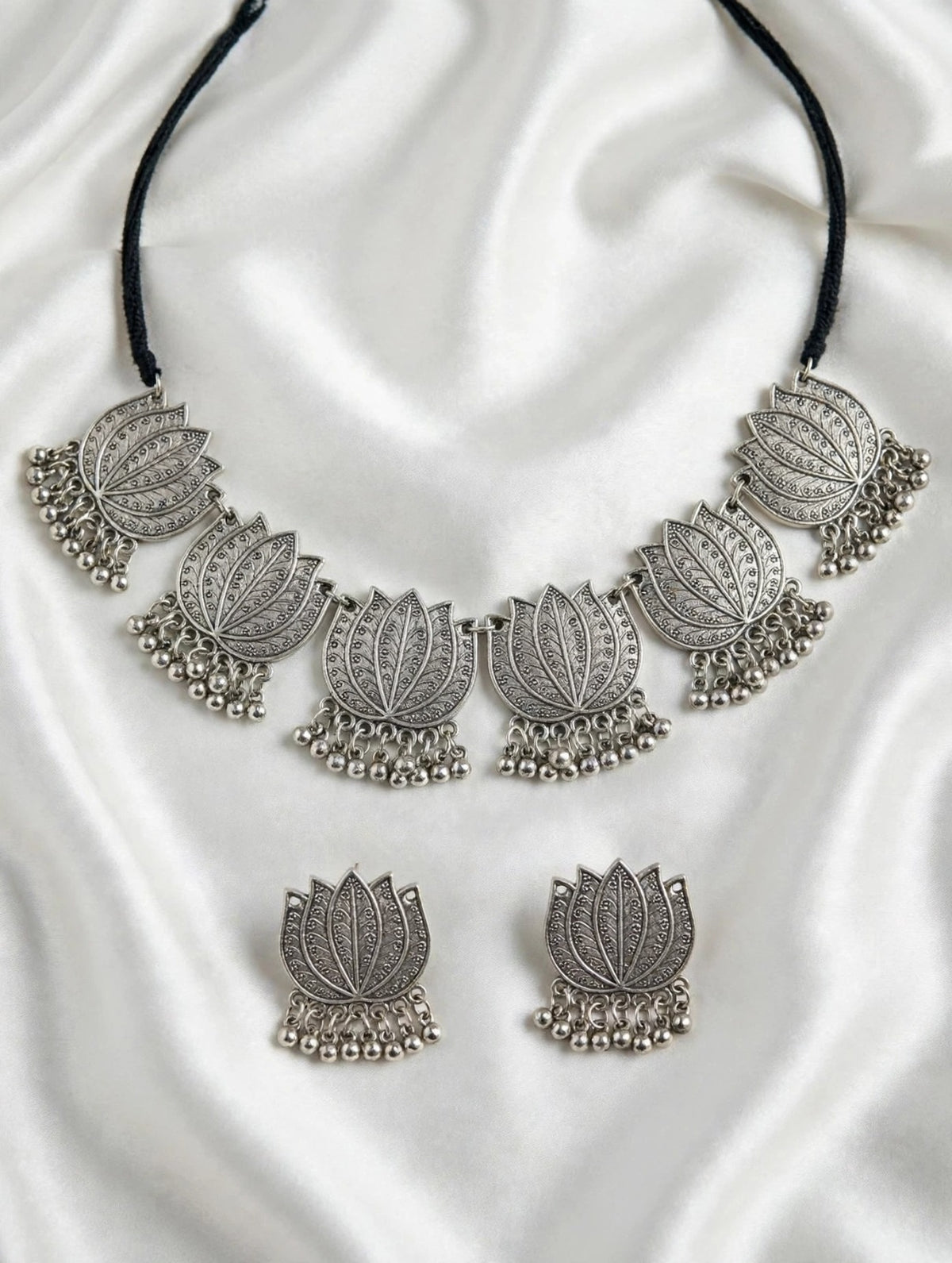 Oxidised lotus necklace with black cord and matching ghungroo-drop earrings arranged on white silk