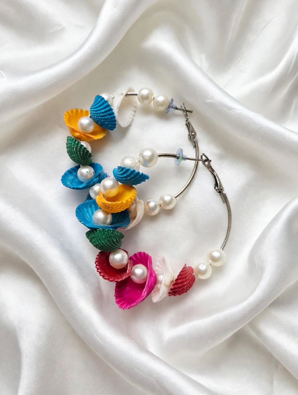 Multicolour shell-shaped hoop earrings with faux pearls and hinged closures on white fabric.