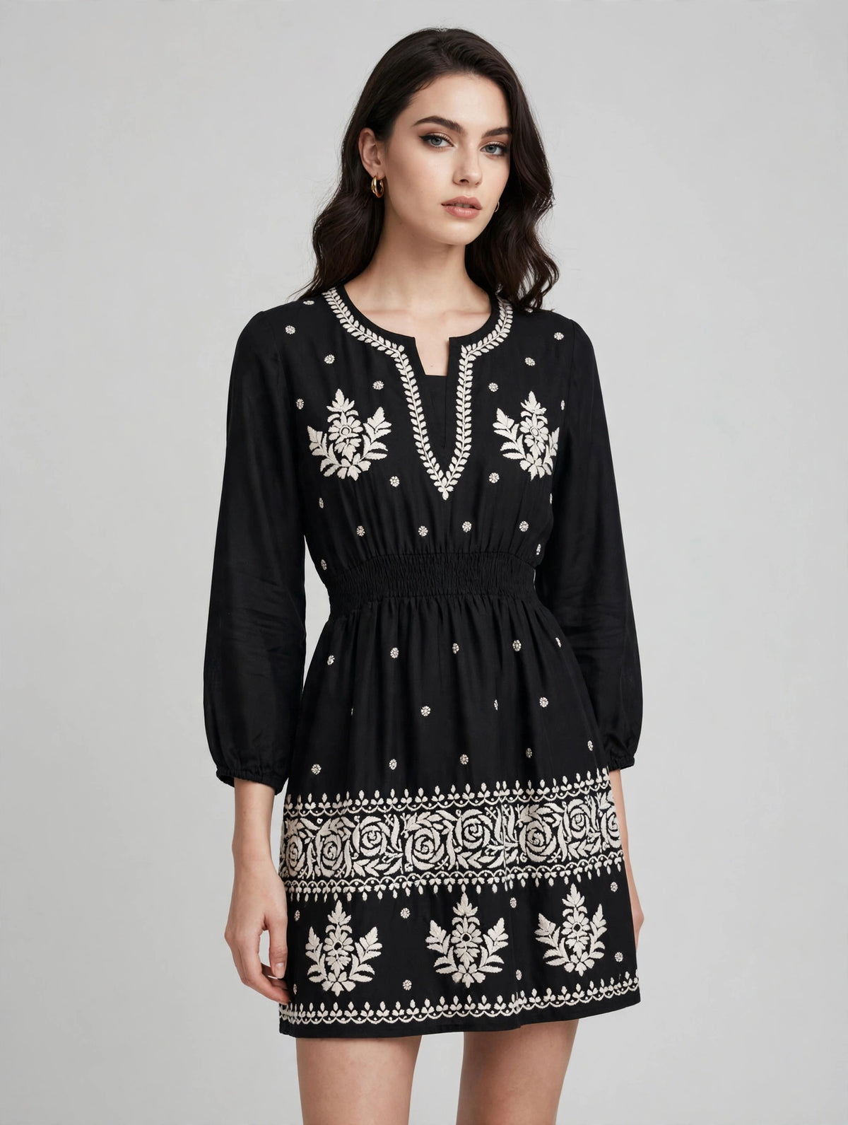 A woman modeling a black long-sleeve mini dress with white embroidered patterns, a split neckline, and a smocked waistband, set against a plain light gray studio background.
