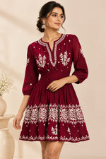 A model wearing a maroon bohemian-style dress with white floral embroidery The dress features a V-neckline, gathered waist, and a tiered skirt with white embroidered patterns.