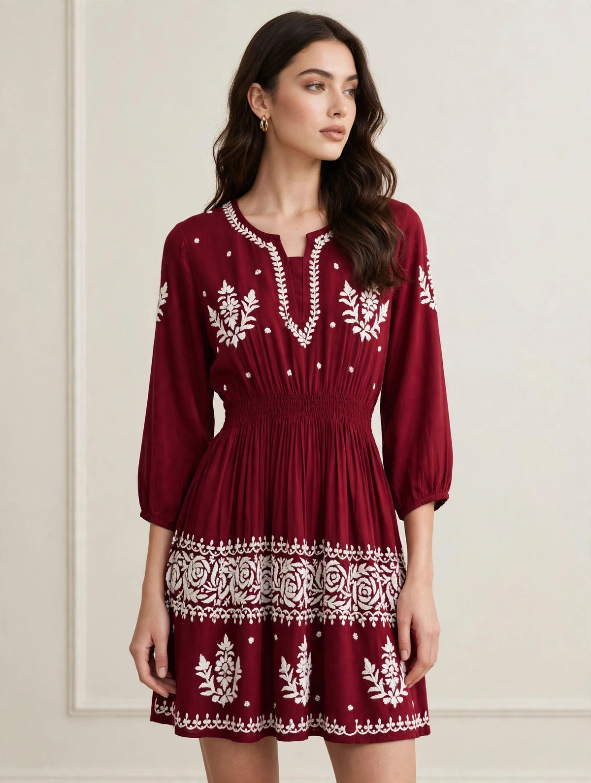 A woman wearing a maroon mini dress with white embroidered patterns, standing against a light beige wall.