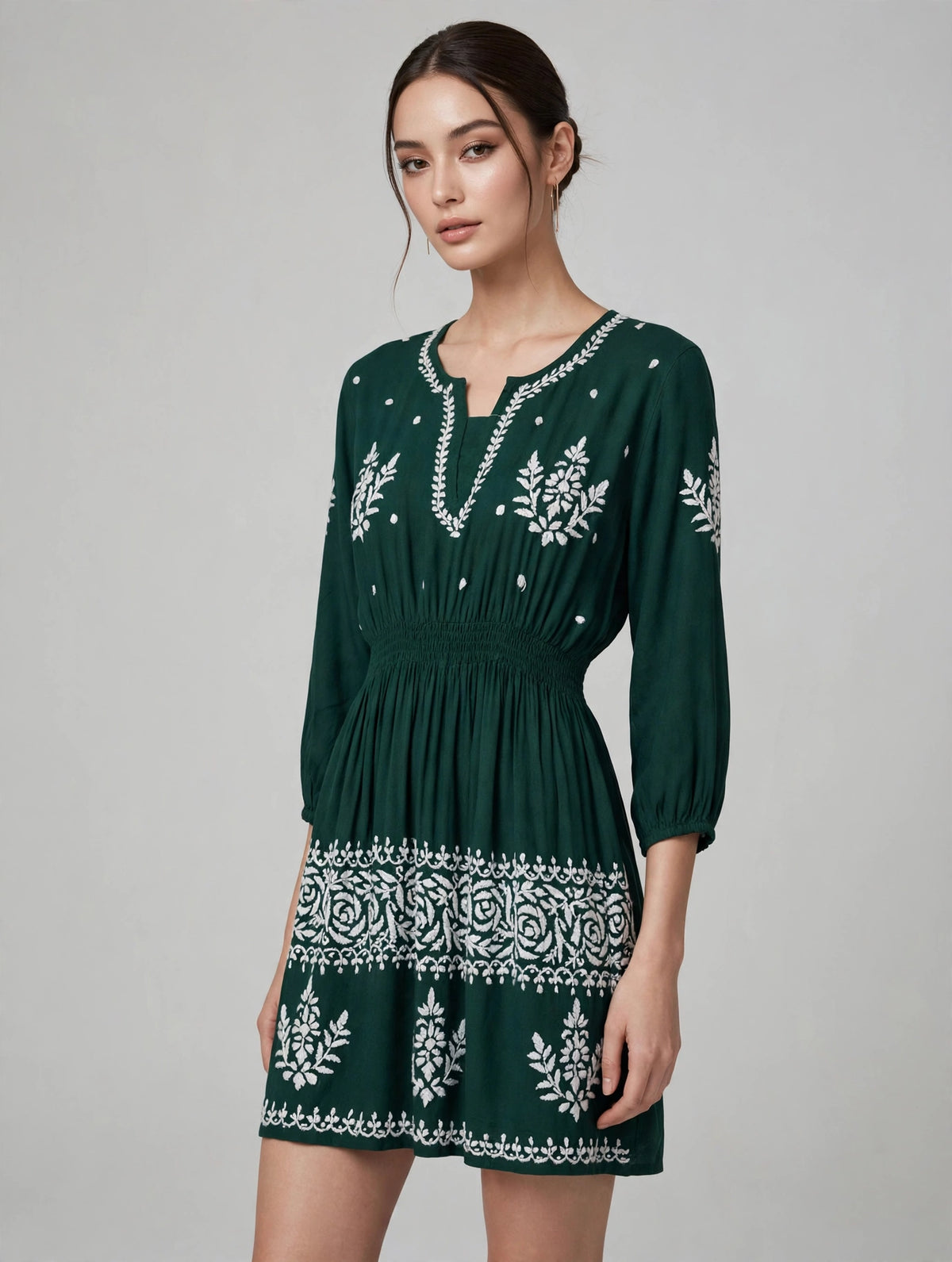 A woman modeling a bottle green mini dress with white embroidered patterns, featuring a split neckline, 3/4 sleeves, and a gathered waist, set against a plain light gray studio background.