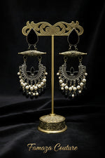 A pair of ornate traditional-style dangle earrings displayed on a black velvet surface. The earrings feature a gold-toned filigree cone bar, silver-toned crescent-shaped metalwork, white pearl beads, and dangling black faceted beads.