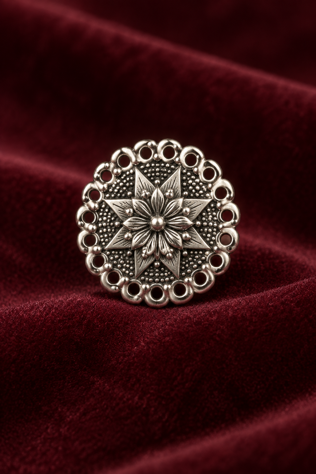 A round silver-toned metal button with an intricate floral starburst pattern and scalloped border, displayed on a draped dark red velvet fabric background.