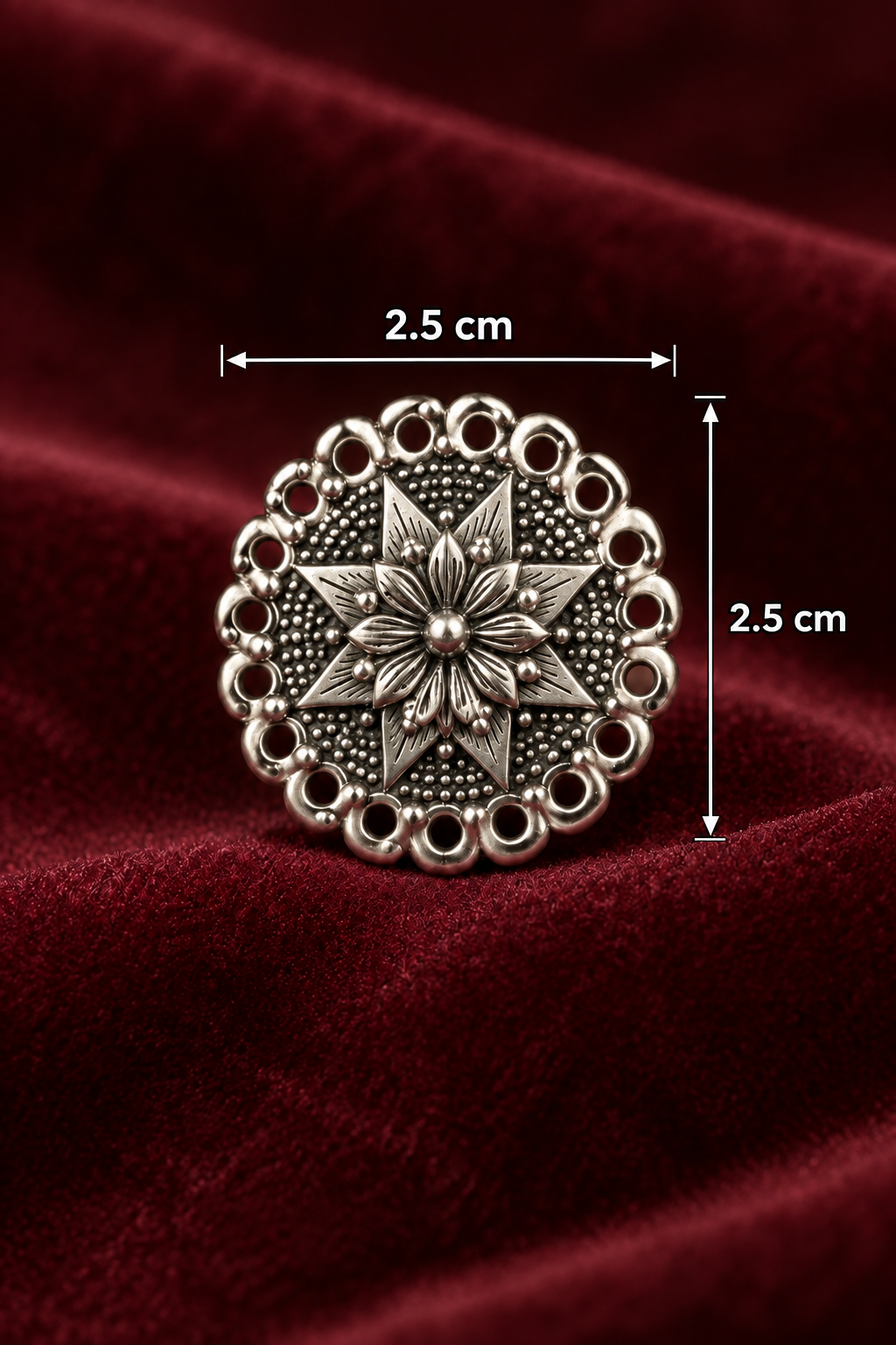 A round silver-toned oxidised stud earrings with an intricate floral starburst pattern and scalloped border, displayed on a draped dark red velvet fabric background.