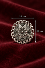 A round silver-toned oxidised stud earrings with an intricate floral starburst pattern and scalloped border, displayed on a draped dark red velvet fabric background.