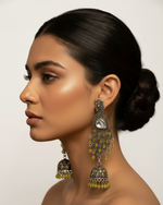 A woman in profile wearing ornate traditional Indian jhumka earrings with silver-toned metalwork, green stones, and clear stones, set against a draped beige fabric background.