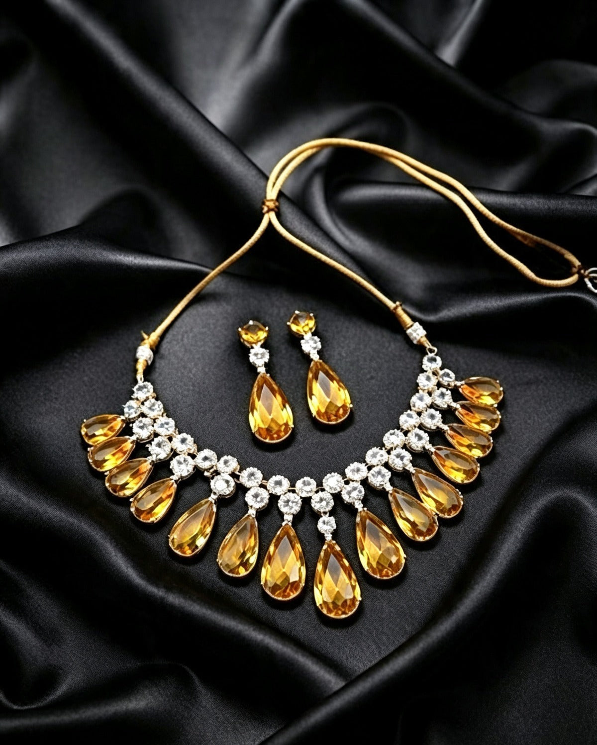A high-angle flat lay view of a micro gold plated yellow crystal and cubic zirconia choker necklace with matching teardrop earrings displayed on a dark satin background.