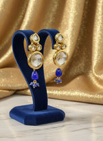 Gold-tone Kundan drop earrings with clear stones and royal blue bead drops on a navy stand.