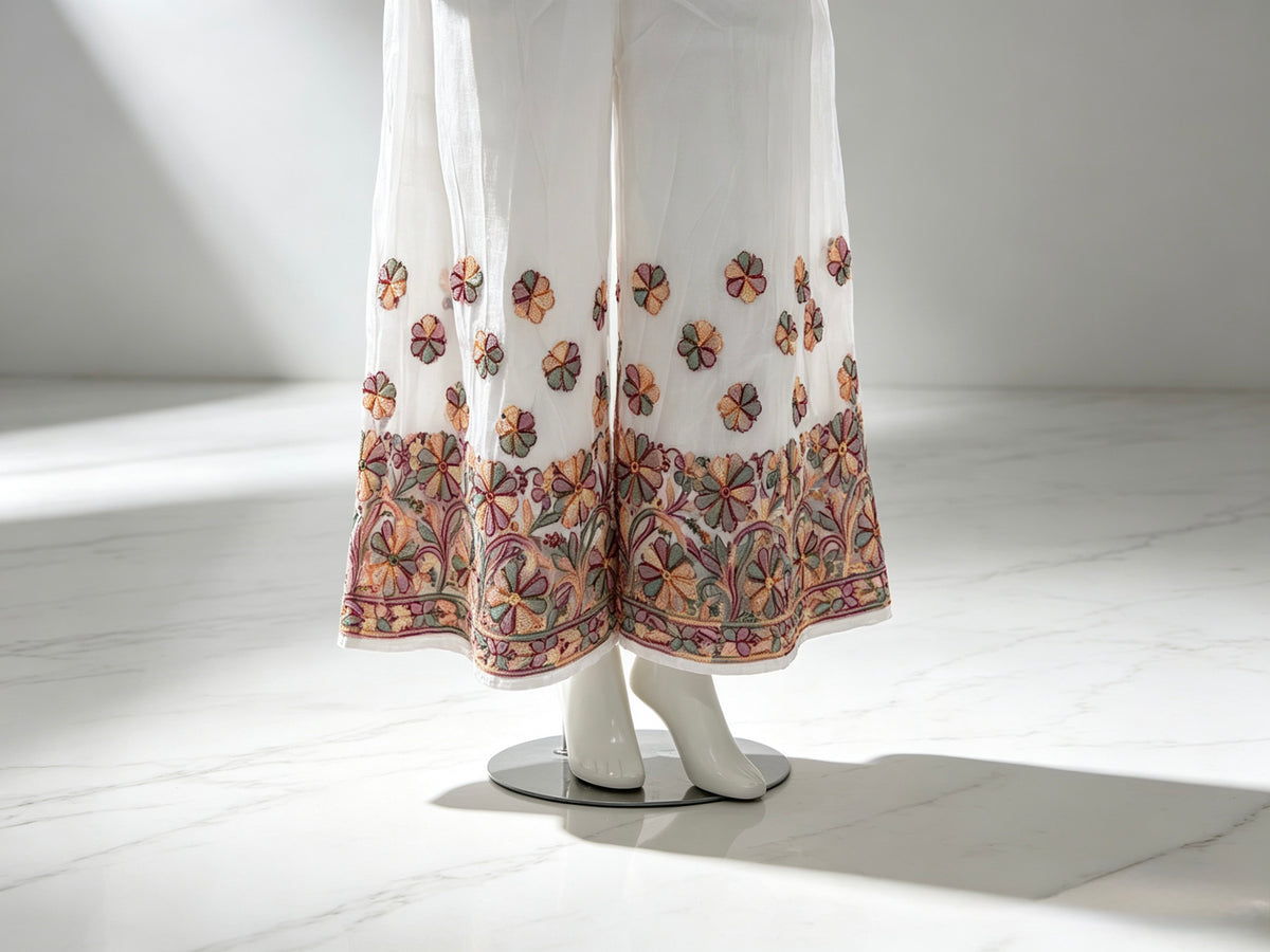 White wide-leg palazzo pants with colourful floral embroidery concentrated along the hem and scattered above the hem, displayed on a white mannequin in a bright, minimalist studio setting.