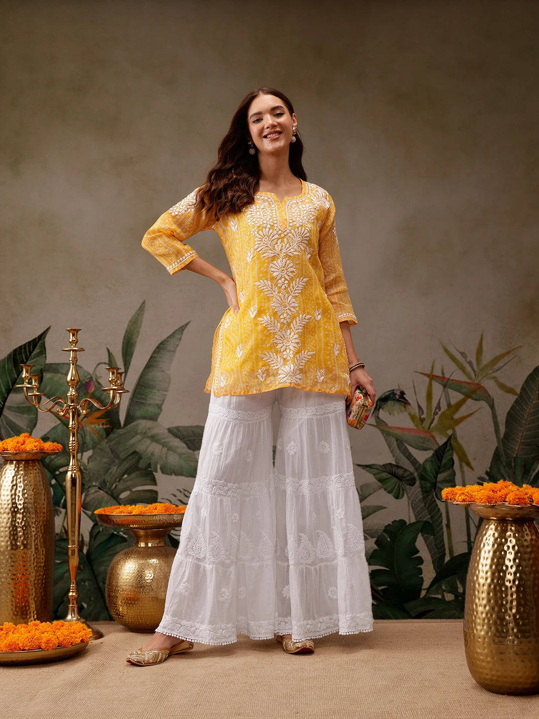 Woman in yellow top and white pants standing in front of decorative vases and plants.