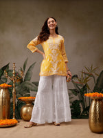 Woman in yellow top and white pants standing in front of decorative vases and plants.