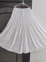 A white tiered skirt with embroidered details and a lace hem, hanging on a hanger against a dark mesh screen and a light wall.