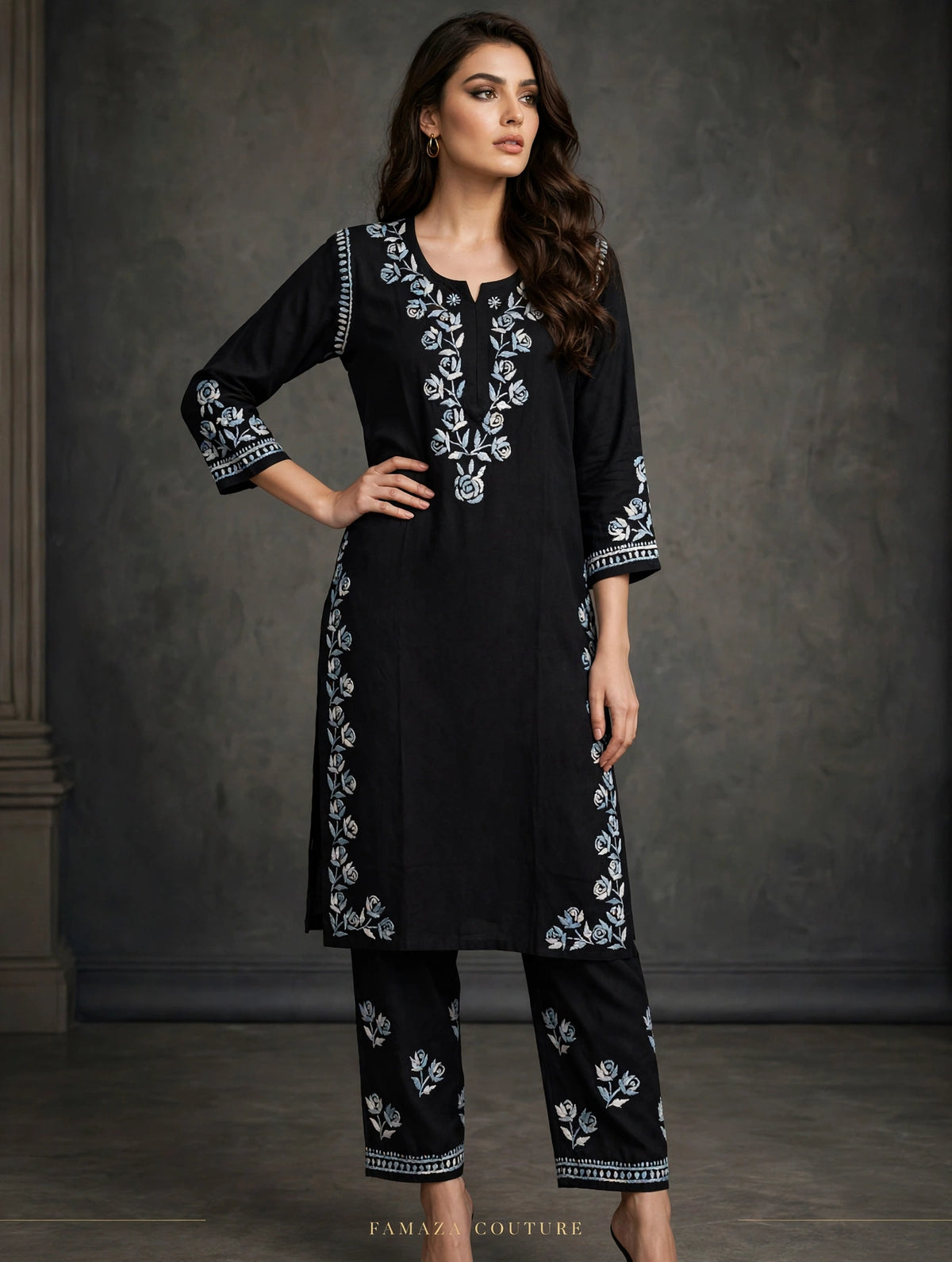 A woman models a black three-piece kurta set with white floral embroidery, featuring a long tunic, cropped pants, and matching belt, set against a dark textured studio backdrop.