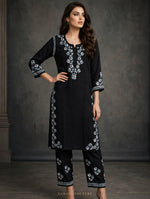A woman models a black three-piece kurta set with white floral embroidery, featuring a long tunic, cropped pants, and matching belt, set against a dark textured studio backdrop.