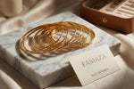 A high-end lifestyle photograph of 12 sleek, micro gold-plated copper bangles with a diamond-cut texture, displayed on a white marble block next to an Aurum Collective Micro Gold Bangles card.