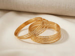 A collection of 12 sleek, micro gold-plated copper bangles with a fine diamond-cut texture, elegantly arranged on a draped cream silk fabric background.
