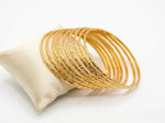 A professional photograph of 12 sleek, diamond-cut textured micro gold-plated copper bangles elegantly resting on a cream-colored silk jewelry cushion against a pure white background.