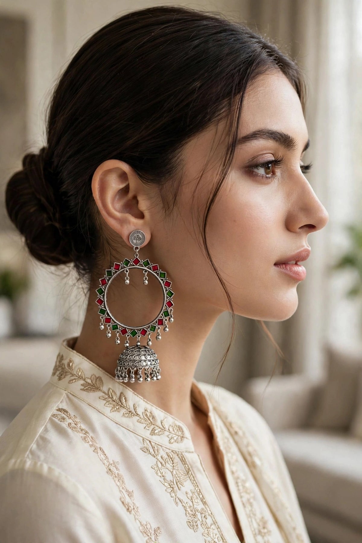 A woman in profile wearing a large silver-toned hoop earring adorned with red and green stones and a dangling bell-shaped charm, paired with a cream embroidered top in a softly lit indoor setting.