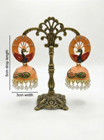 5.5cm orange Meenakari peacock jhumkas with hand-painted enamel and white pearl fringe on a professional stand.