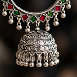 A close-up of an ornate silver-toned traditional necklace with a large bell-shaped pendant, decorated with intricate scrollwork and dangling silver beads, featuring red and green square accents at the top.