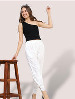 Woman in black top and white pants posing next to a wooden stool against a light background