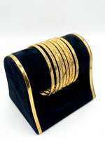 A set of eight micro gold-plated copper bangles with an intricate twisted rope and wavy vine texture, displayed on a posh black velvet and gold-trimmed jewelry stand against a white background.