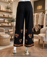 Black wide-leg cropped pants with orange and beige floral embroidery and a decorative border, displayed on a mannequin in a boutique setting.