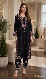 A woman models a black three-piece kurta set with pink floral embroidery in an elegant indoor boutique setting. The set includes a long kurta with elbow-length sleeves, matching trousers, and a coordinating belt.