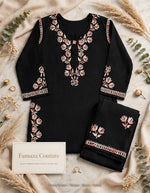 A black embroidered kurta set with matching pants, laid flat on a beige fabric background with decorative dried flowers and greenery. The kurta has pink and beige floral embroidery around the neckline and sleeves, with a small pink bow at the collar.