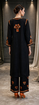 A woman stands with her back to the camera wearing a black traditional outfit with orange floral embroidery on the neckline, sleeves, and pant hems, paired with orange high heels in an indoor setting.