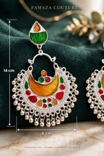 A close-up of a silver crescent-shaped earring with colorful enamel details in green, orange, red, and yellow, displayed on a dark green velvet surface with other matching earrings partially visible.