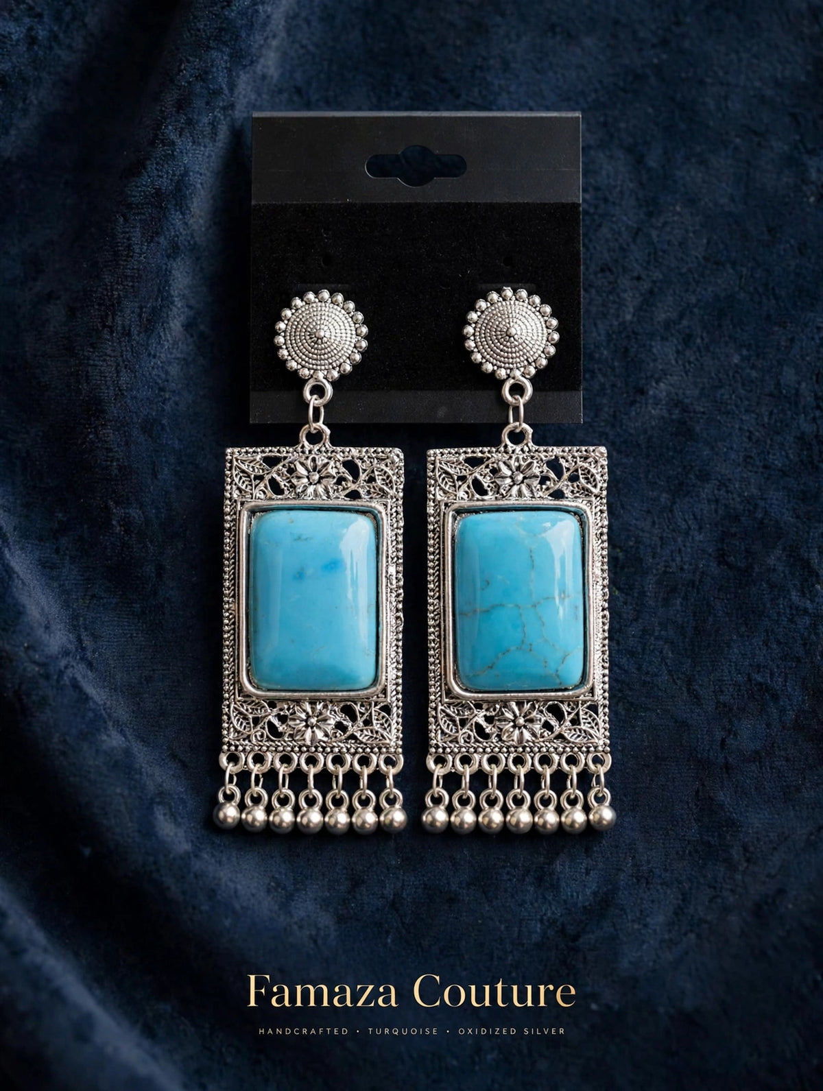 A pair of ornate silver-toned drop earrings with rectangular turquoise blue stone centers and dangling bead chains, displayed on a black card against a dark blue textured fabric background. The brand name 'Famaza Couture' is printed at the bottom.