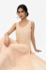 A woman modelling a sleeveless peach-pink straight kurti of viscose fabric with intricate white floral embroidery on the bodice and scattered embroidered motifs on the flowing skirt, set against a plain white background.