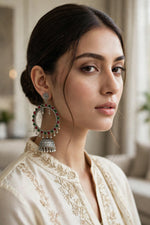 A close-up portrait of a woman wearing a large ornate silver-toned jhumka earring with red and green stone accents, paired with a cream embroidered traditional top in a softly lit indoor setting.