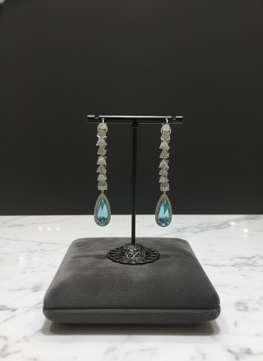 A pair of silver finish American diamond Jhumka bali earrings with green and clear stones on a stand.