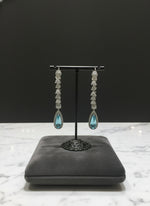 A pair of silver finish American diamond Jhumka bali earrings with green and clear stones on a stand.