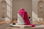A Hot pink shawl with floral patterns draped over a mannequin next to a black handbag on a small table against a marble wall.