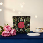 A black floral handcrafted tote bag with glass beads embroidered on a white silk base, displayed on a wooden surface with a greenery background.