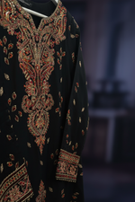 A close-up of a black traditional embroidered garment featuring ornate red and gold paisley patterns on the neckline, sleeve cuff, and hem.