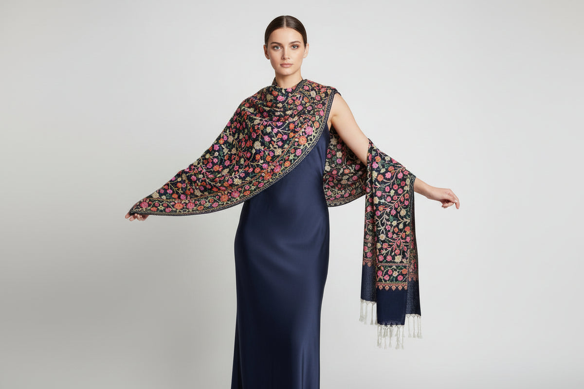 A black scarf with elaborate pink and green floral aari embroidery and tassels on the ends, displayed on a dark background.