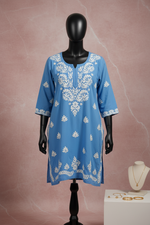 A blue long-sleeve short tunic top with white floral and leaf embroidery, hanging on a wooden hanger against a white draped fabric background.
