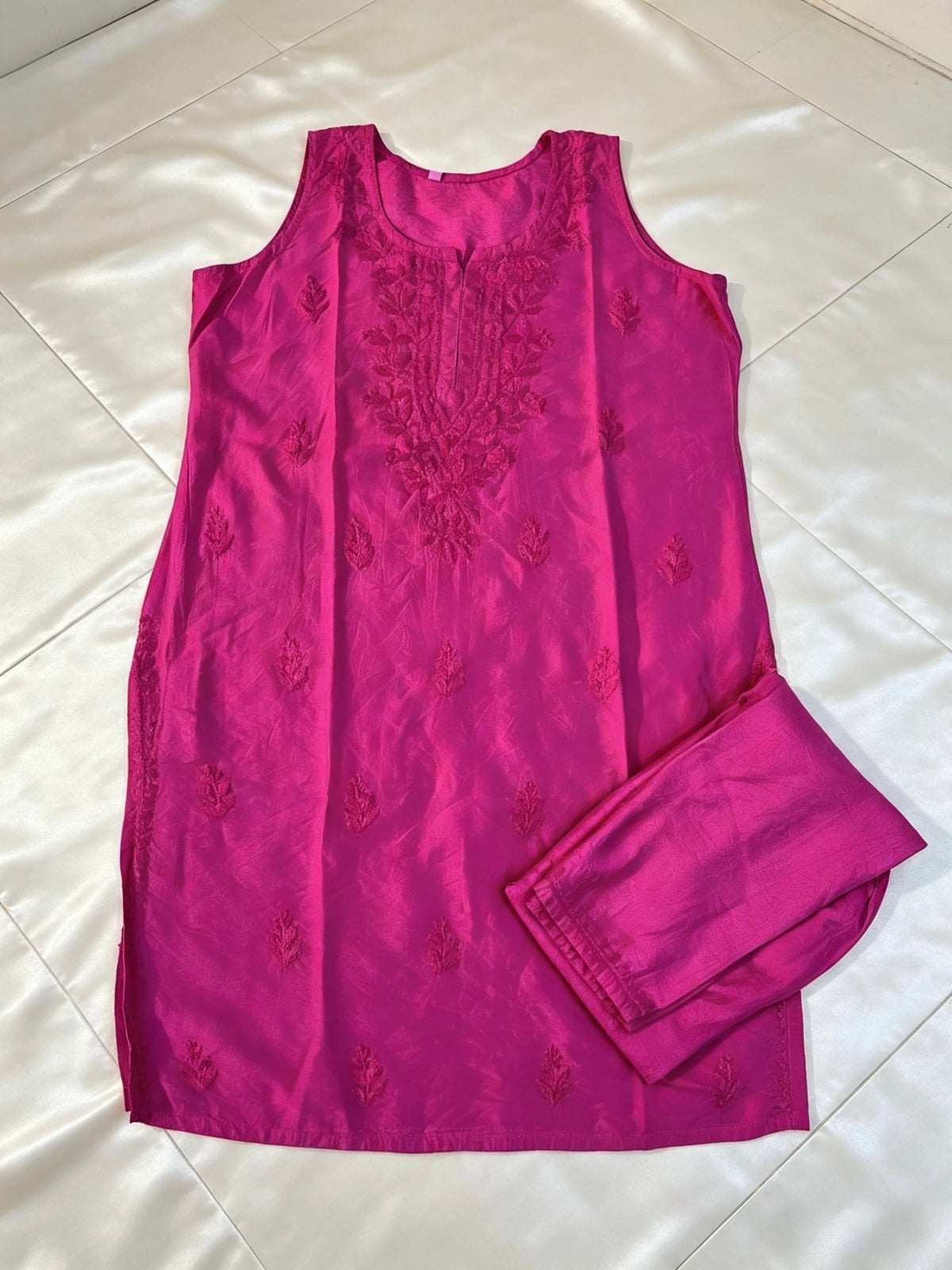 A bright pink sleeveless kurta with matching pants, laid flat on a white satin fabric background. The kurta features subtle tonal floral embroidery and a round neckline.