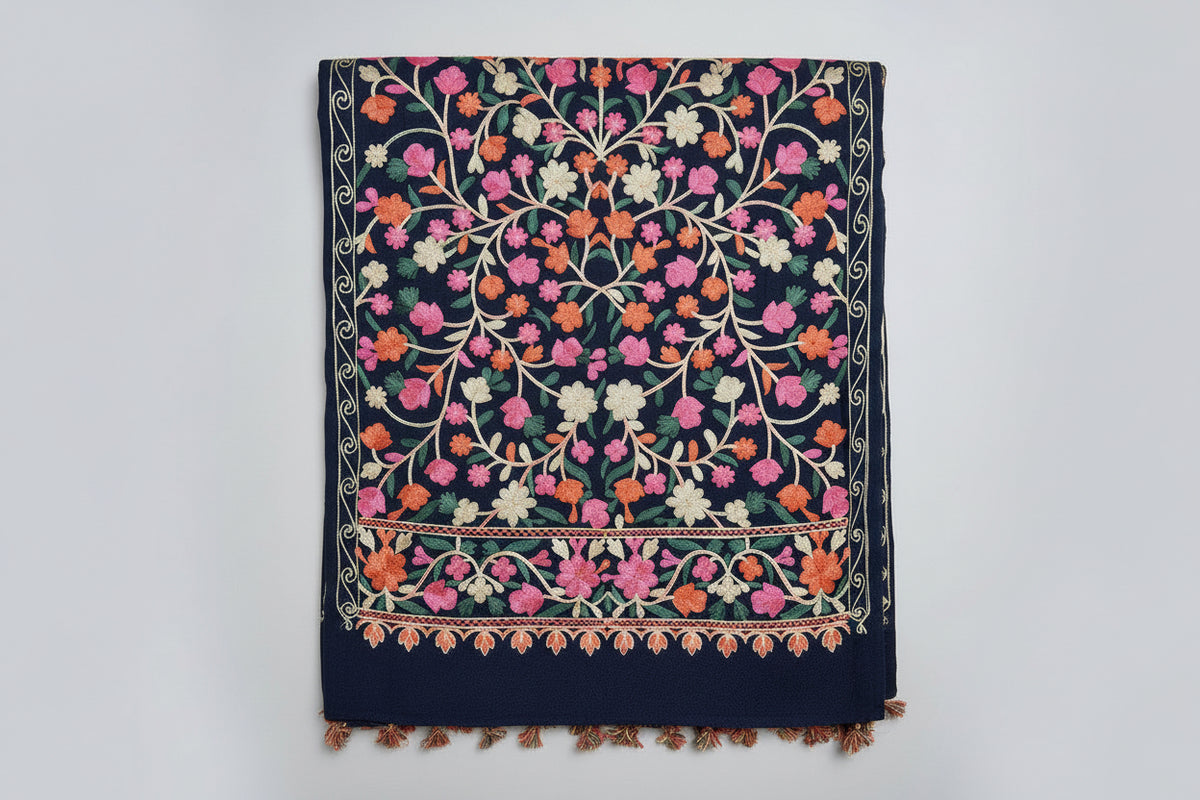 A close-up black scarf with elaborate pink and green floral aari embroidery and tassels on the ends, displayed on a dark background.