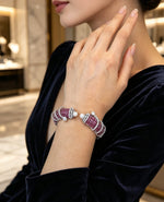 A close-up lifestyle shot of a model wearing a multi-strand bracelet with red gemstone-like beads, white pearl-like accents, and silver-toned metal links. The model is wearing a dark navy velvet top, and the background shows a softly blurred luxury interior.