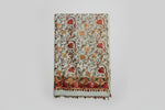 A close-up of White base colour Kashmiri embroidered pashmina scarf with maple leaf motifs and tassels on the ends, displayed on a wooden surface.