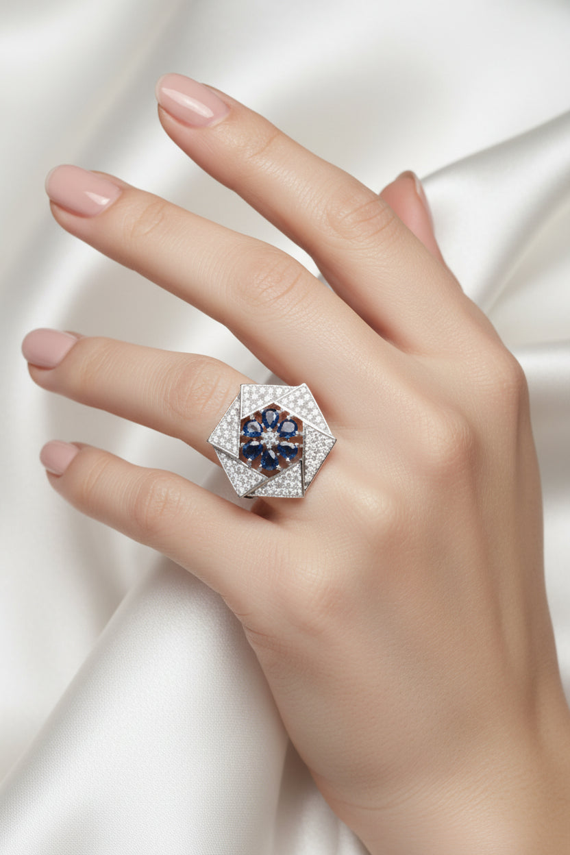 A close-up of a hand holding a geometric silver ring featuring a blue sapphire flower motif in the centre and pavé-set clear stones on layered triangular petals, set against a textured blue fabric background.